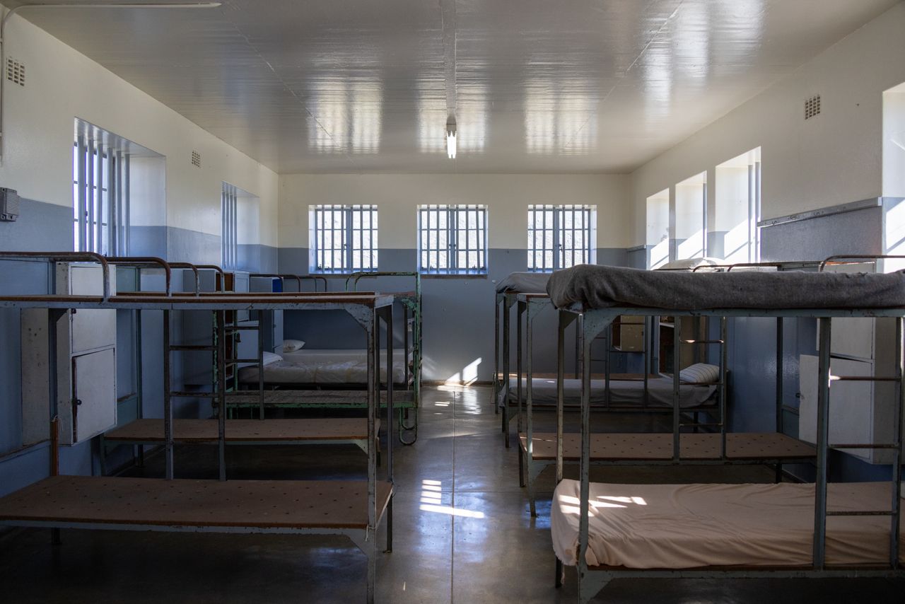 A vacant institutional dorm room with metal bunk beds and barred windows, lit by natural light.